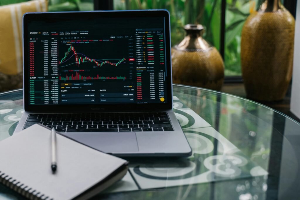 pexels-photo-14751274-14751274 Laptop displaying cryptocurrency stocks and graphs on a glass table with a notepad.